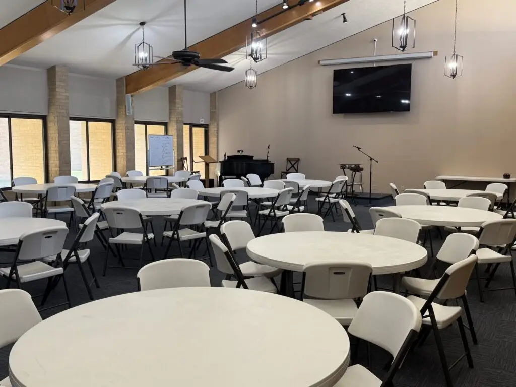 The fellowship hall set with round tables and folding chairs, exposed timber beams and pendant lanterns overhead, a wall-mounted screen and a small stage with a piano at the far end.