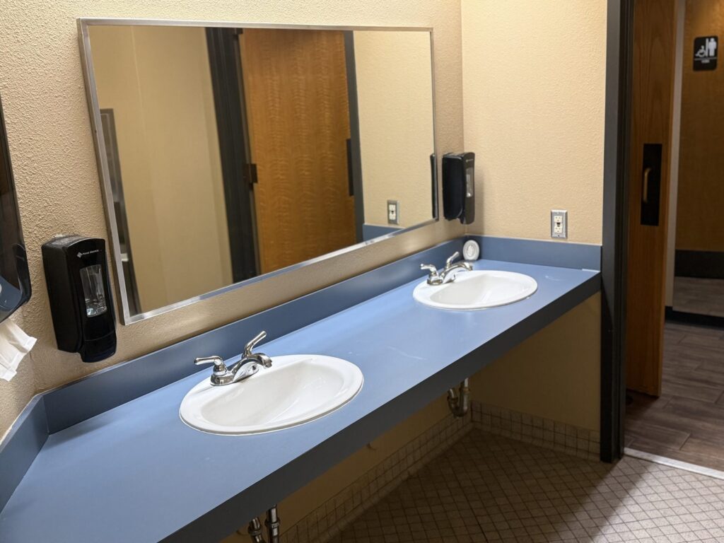 The men's restroom: two basins set in a long blue laminate counter beneath a full-width mirror, with soap dispensers on a tan wall and a tiled floor.